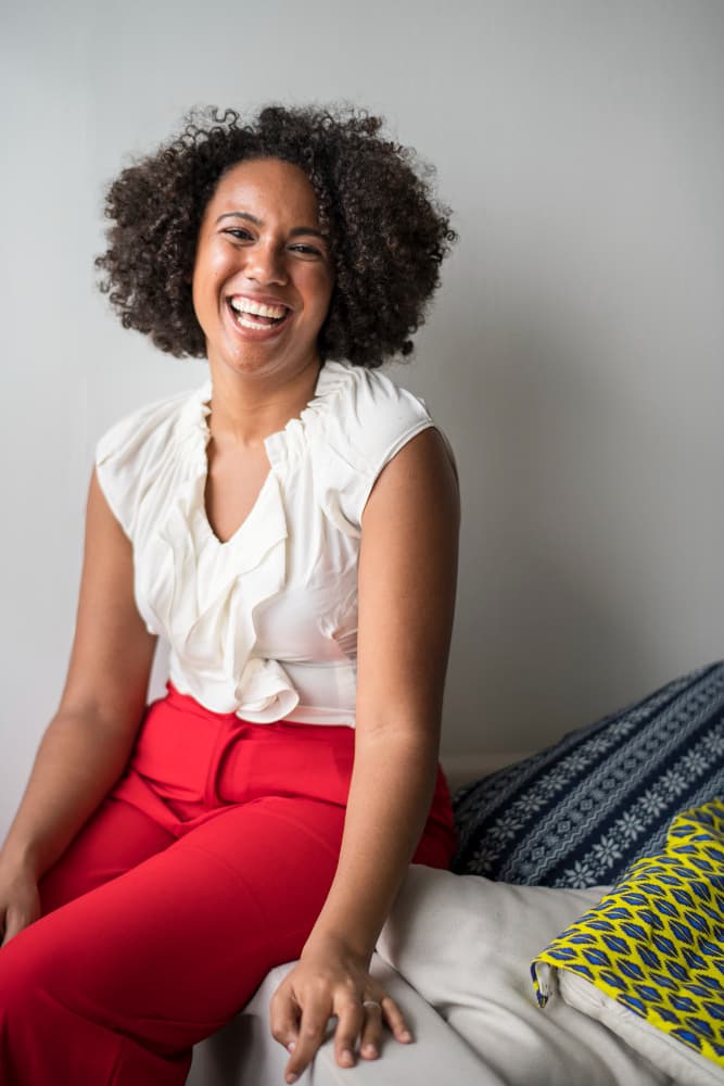 Mulher negra sorridente com cabelo crespo, blusa branca e calça vermelha, sentada em um sofá.