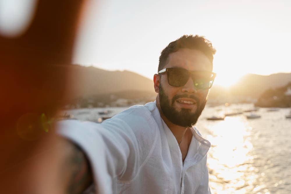 Homem barbudo de óculos escuros tira selfie no litoral durante o pôr do sol.