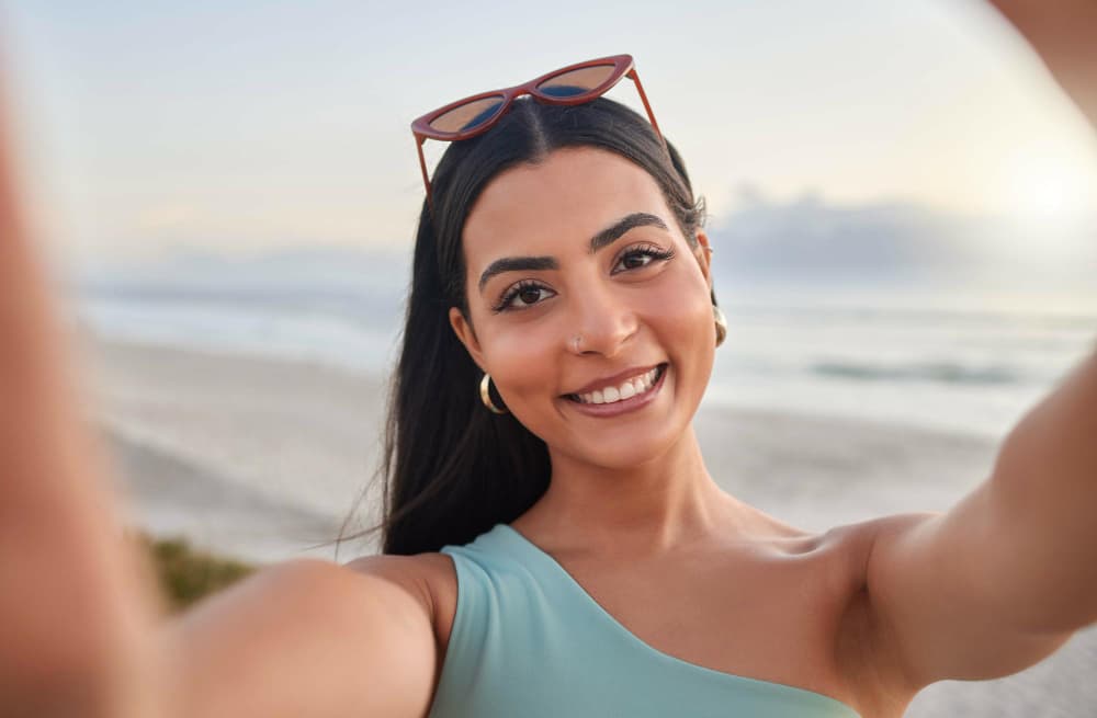 Mulher sorridente tirando selfie na praia com óculos de sol e mar ao fundo.