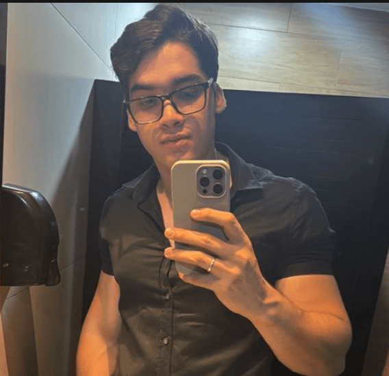 Homem jovem de óculos tira selfie no espelho vestindo camisa preta de botões.