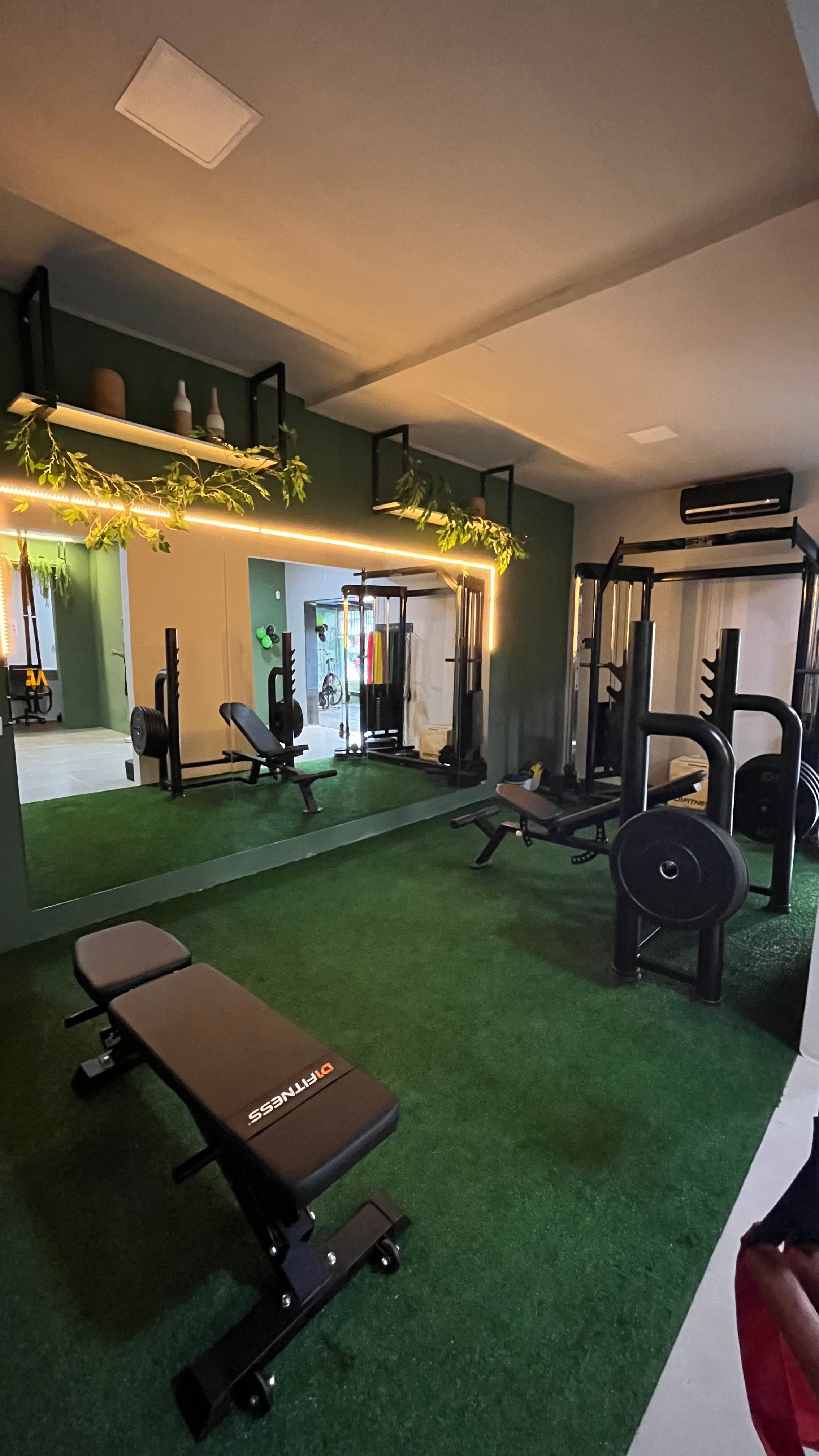 Academia com grama sintética verde, bancos de musculação, espelhos grandes e iluminação quente e moderna.