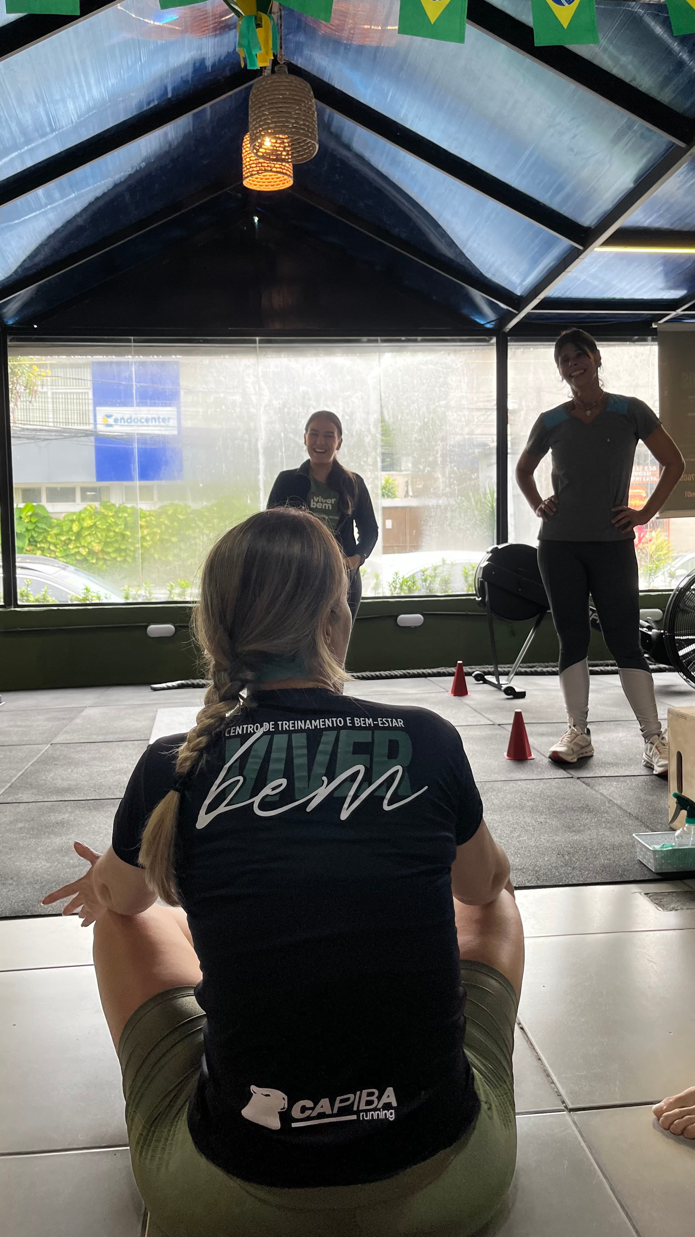 Mulher sentada de costas em academia observando duas mulheres sorridentes em um centro de treinamento.