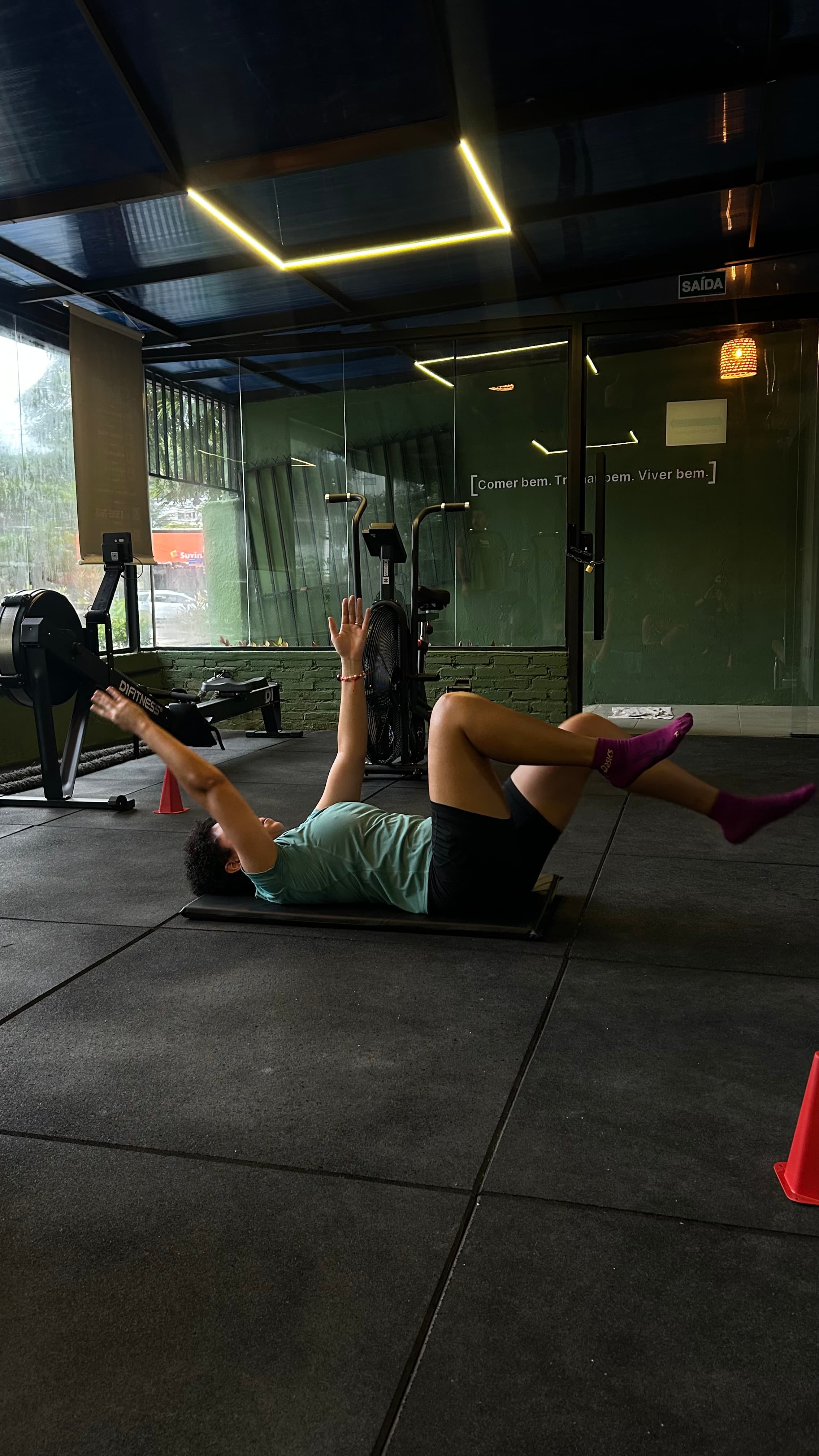 Pessoa deitada em colchonete preto realizando exercícios abdominais em uma academia com equipamentos ao fundo.